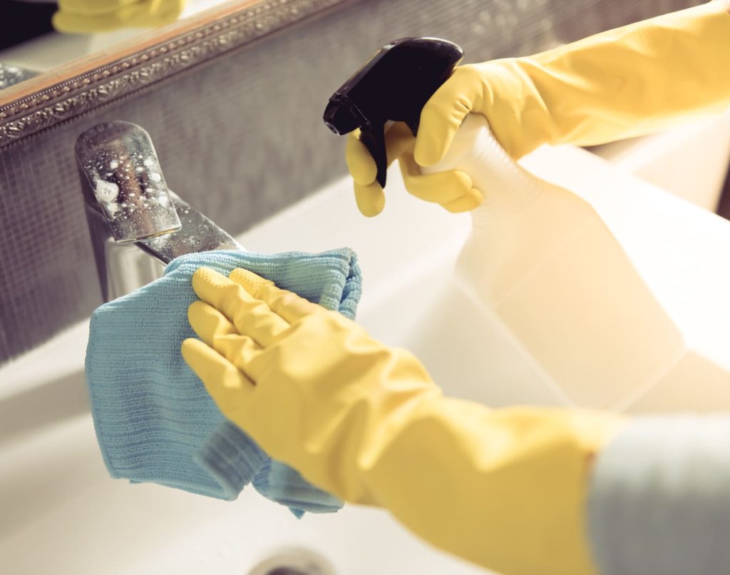 Manos con guantes amarillos limpiando un grifo con un paño y un spray.